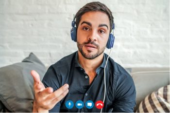 A camera view of a young man wearing headphones who appears to be on a virtual meeting, who looks like is is asking a question